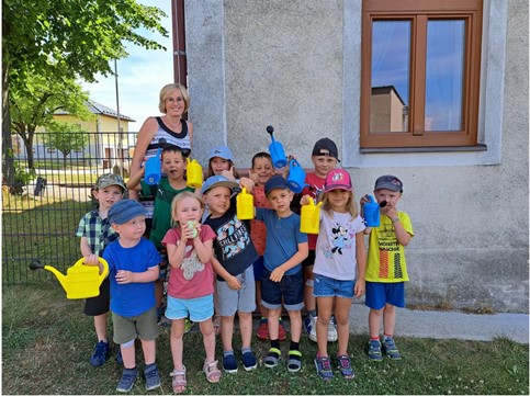 "Regenwasser-Be-Halte-Region": Eine 11-köpfige Kindergartengruppe gemeinsam mit ihrer Aufsichtsperson vor einer Hauswand. Einige der Kinder halten gelbe Gießkannen in die Kamera.