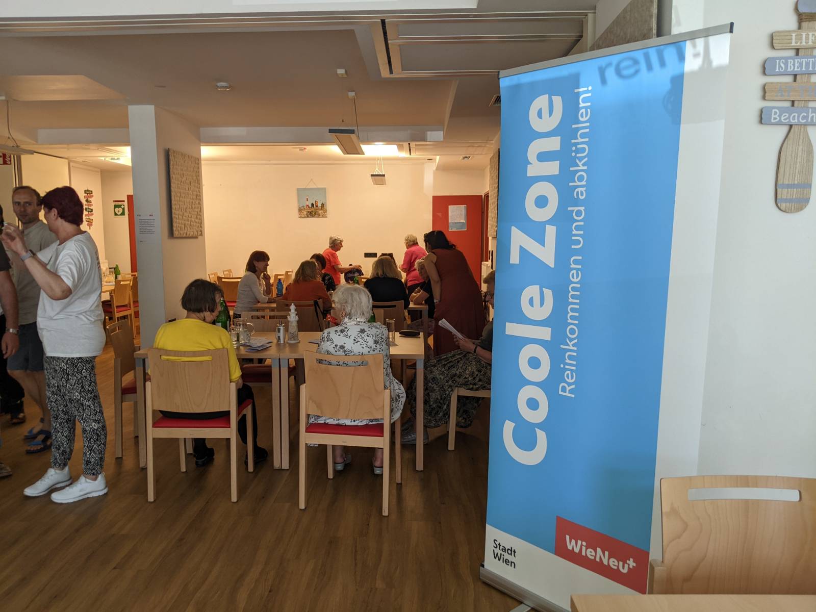 "Coole Zonen - reinkommen und abkühlen!": Man sieht in einen Innenraum hinein, in dem mehrere Menschen auf Stühlen und an Tischen sitzen. Rechts im Bild steht ein Roll-Up, auf dem in großen Buchstaben "Coole Zone - Reinkommen und abkühlen!" steht.