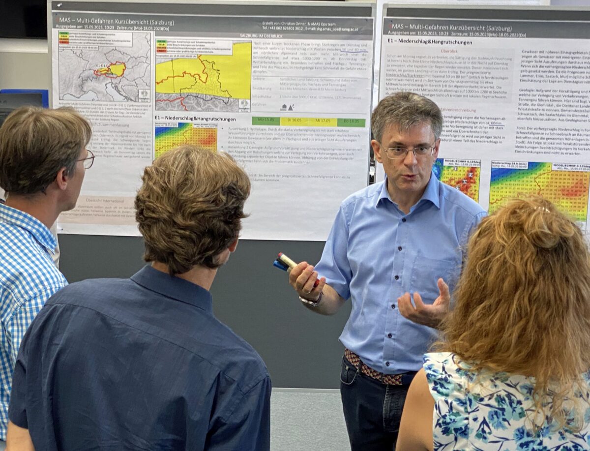 Vier Personen stehen vor einer Wand mit wissenschaftlichen Postern, eine Person hält einen Zeigestab und erklärt etwas.