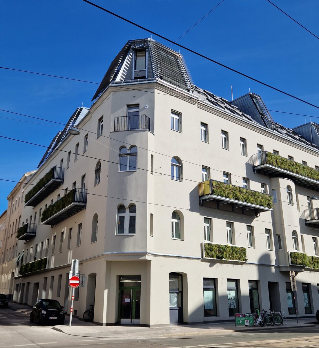 Eckgebäude im Gründerzeitstil mit Mansarddach, mehreren Balkonen mit Pflanzen und hell verputzter Fassade.