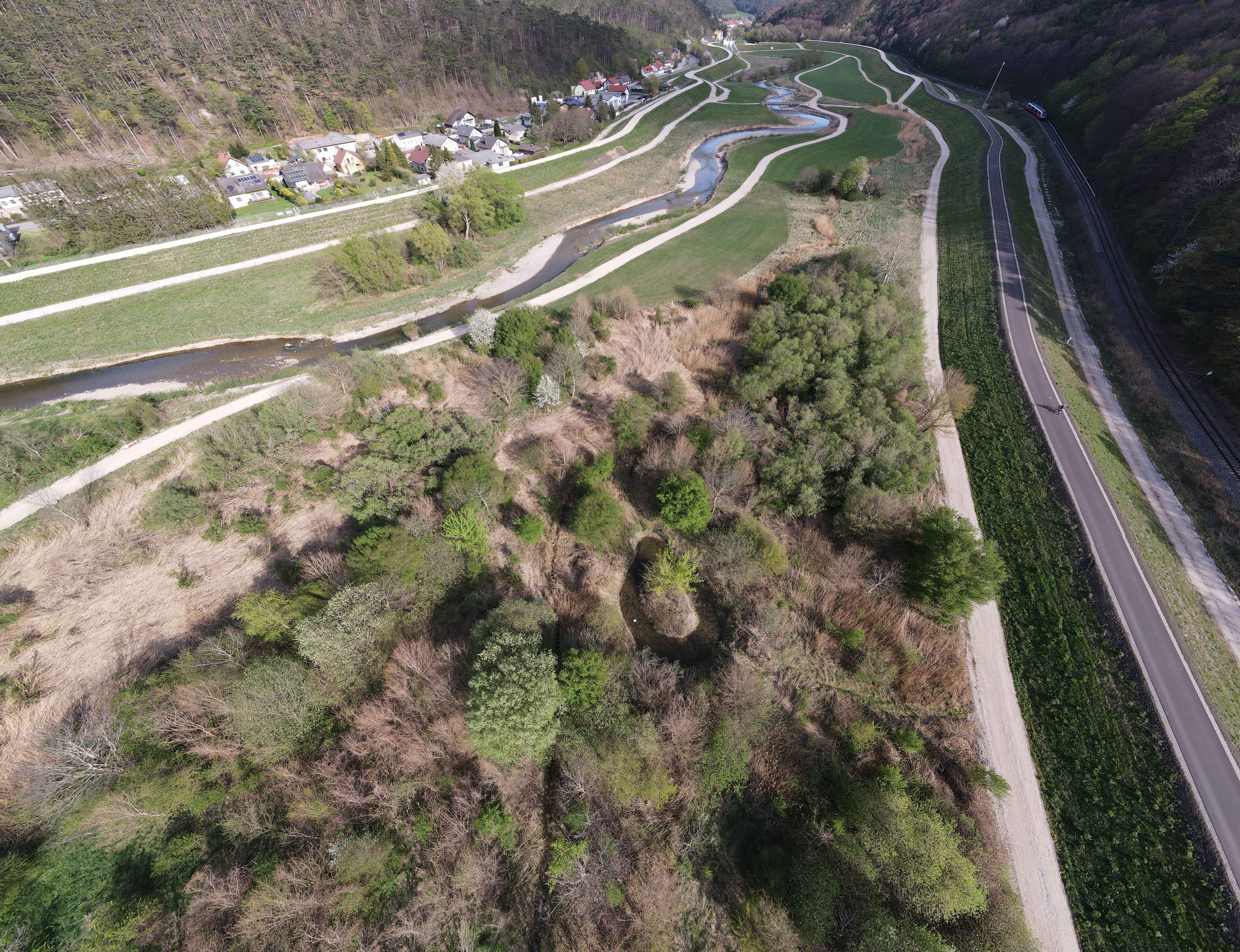 Luftaufnahme eines Flusstals mit gewundenem Fluss, angrenzenden Wiesen, Waldflächen und einer Straße rechts.