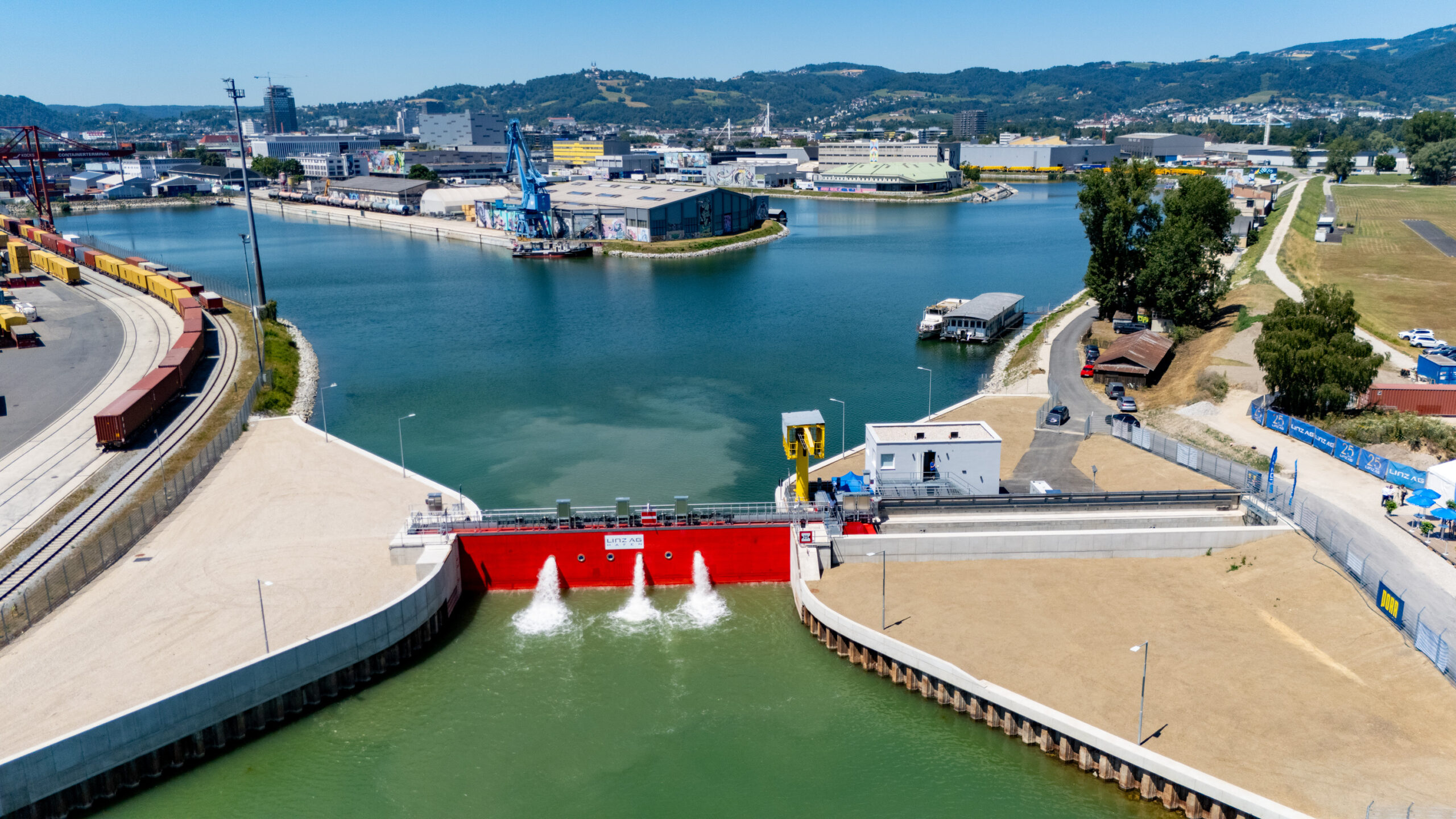 Luftaufnahme des Hochwasserschutzes vom Handelshafen Linz mit rotem Schleusentor, angrenzenden Lagerflächen und Industriegebäuden im Hintergrund.